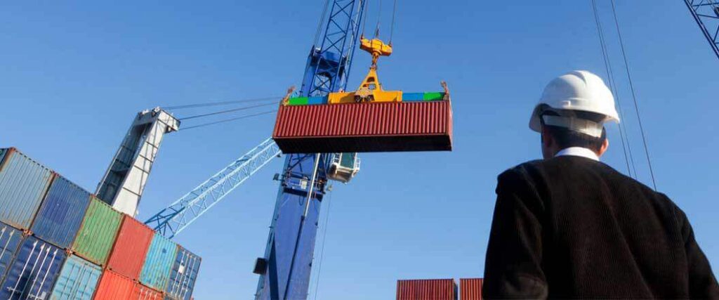 Person in a hard hat looking up a crane holding a shipping container. 