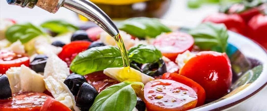 Olive oil being poured onto a bed of sliced tomatoes, olives, and basil leaves.