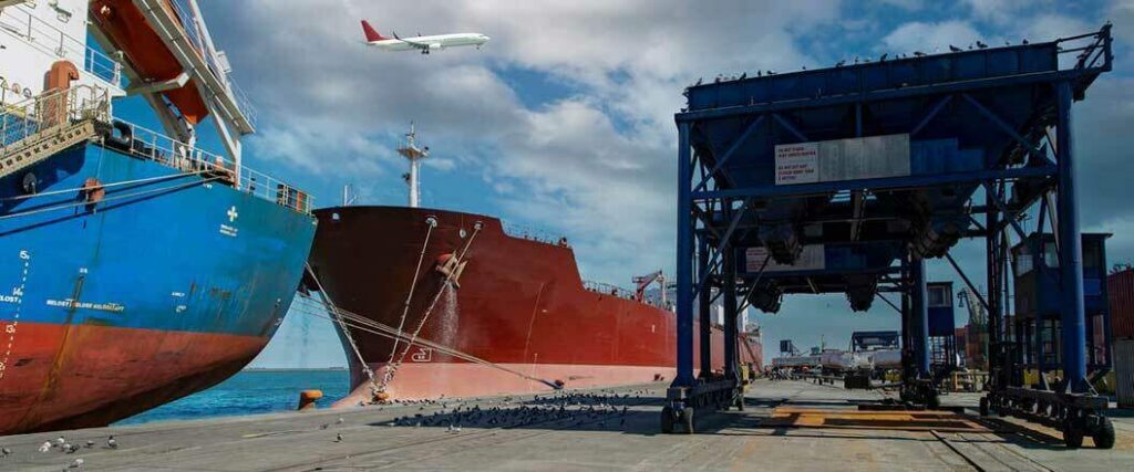 Two cargo vessels docked at port with a plane flying overhead.