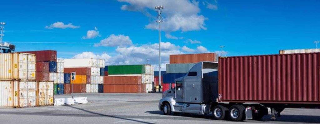 A container truck at a dock approaching stacks of containers waiting to be loaded.